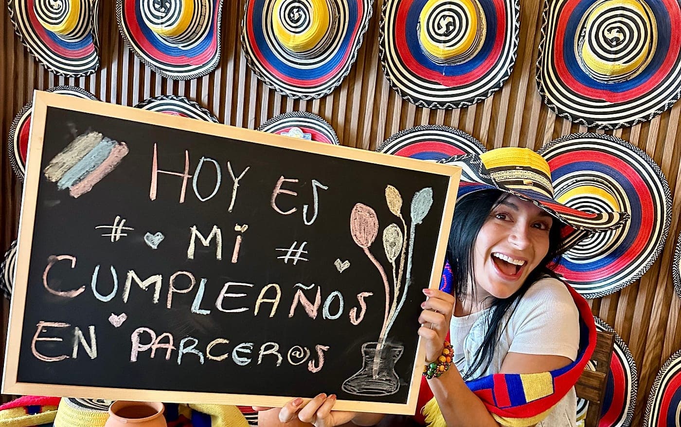 Una persona con sombrero vueltiao sostiene, sonriendo, un pizarrón que dice «Hoy es mi cumpleaños en Parceros», frente a una pared cubierta de sombreros vueltiaos