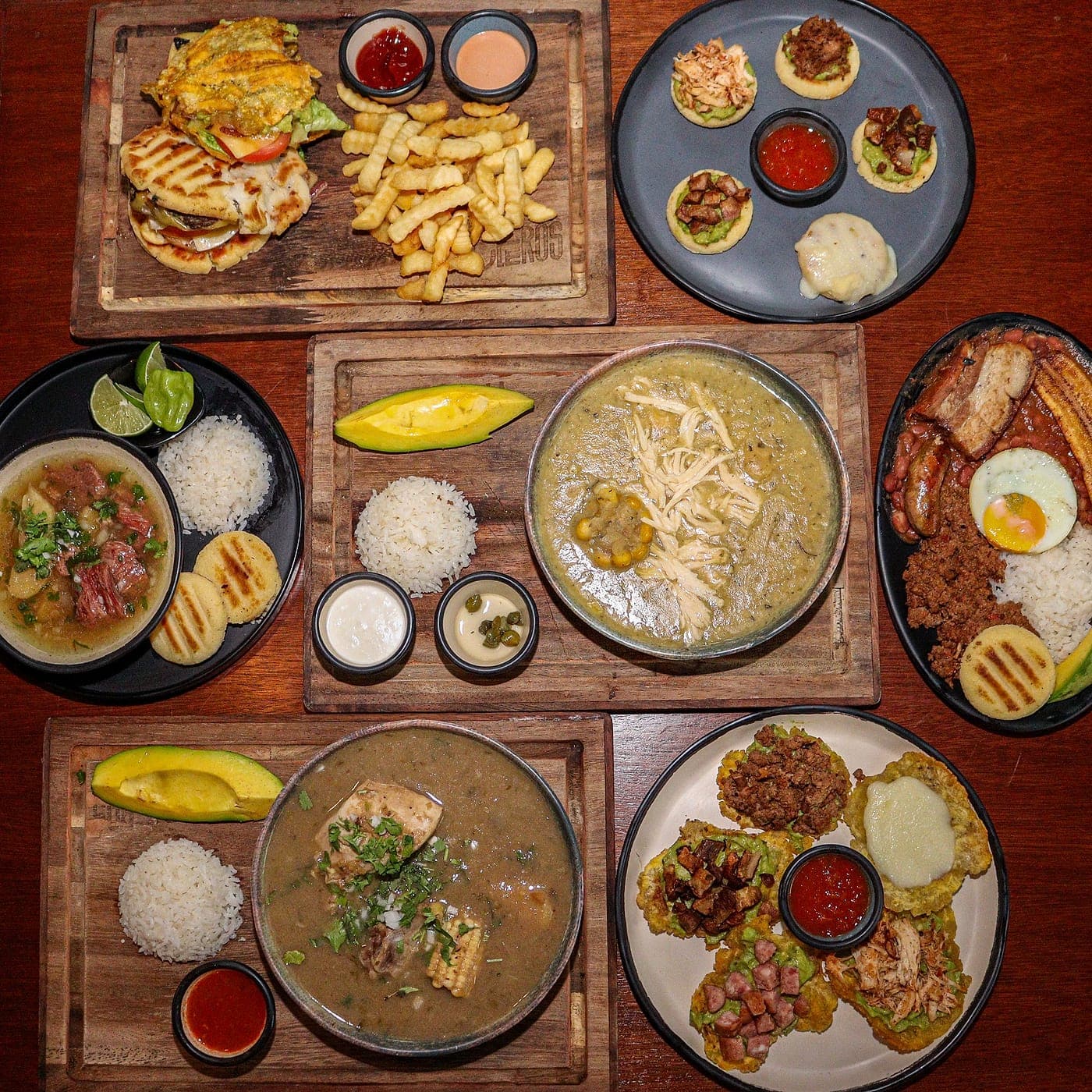 Vista cenital de una mesa entera de Parceros cubierta de platos: hamburguesas, sopas, patacones, bandeja paisa, arroces y salsas