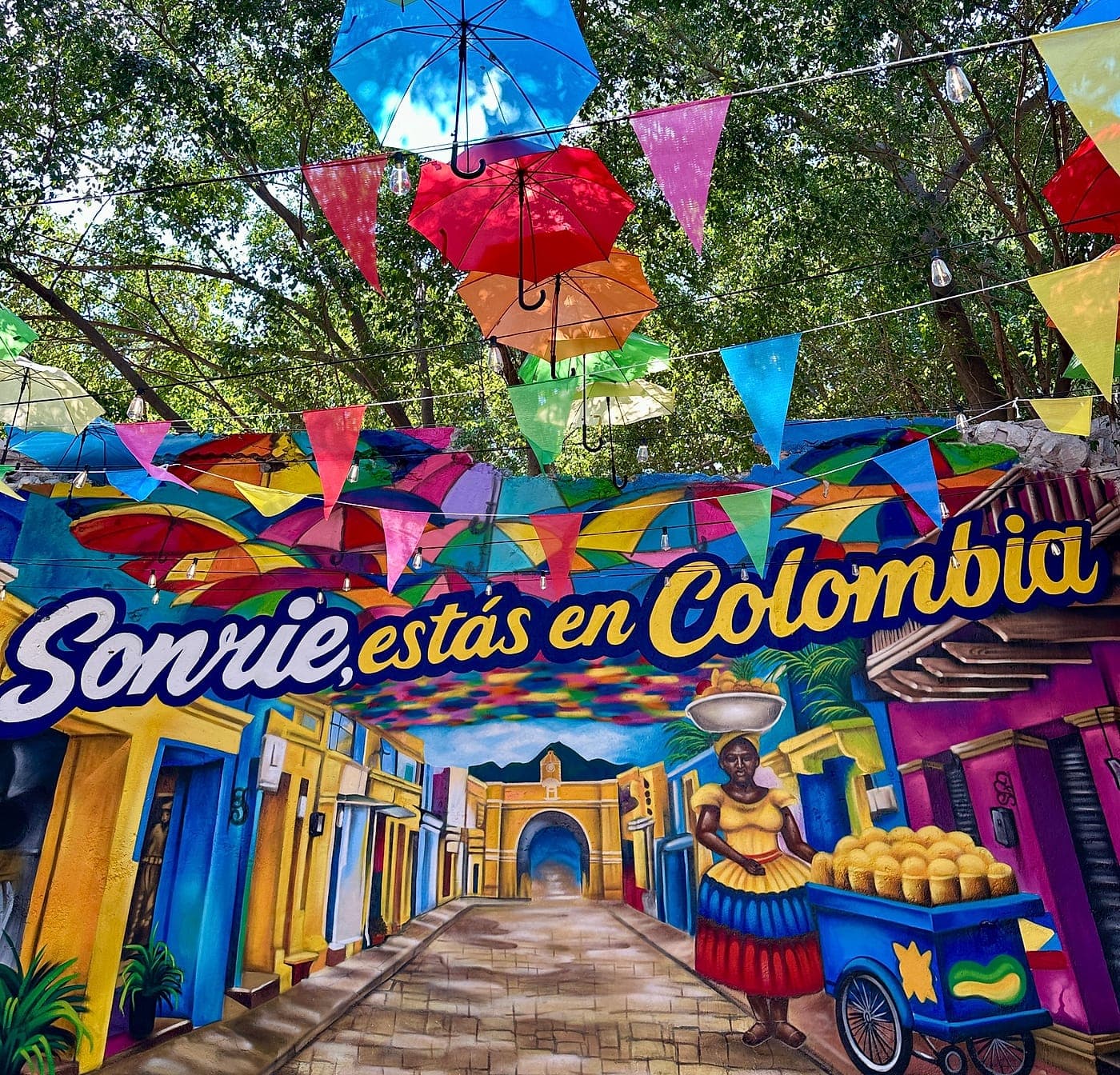 Mural «Sonríe, estás en Colombia» a plena luz: una calle colonial de casas de colores con una palenquera y su carreta de mangos, bajo sombrillas y banderines