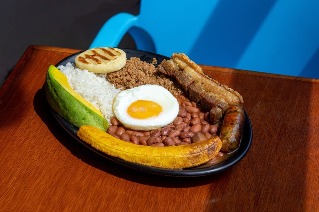 Bandeja Paisa Personal: El plato que mejor representa a Colombia. Una explosión de sabores dulces, salados y crocantes en una sola bandeja. Frijoles, chicharrón, chorizo, carne, huevo estrellado, aguacate, arroz, plátano y arepa